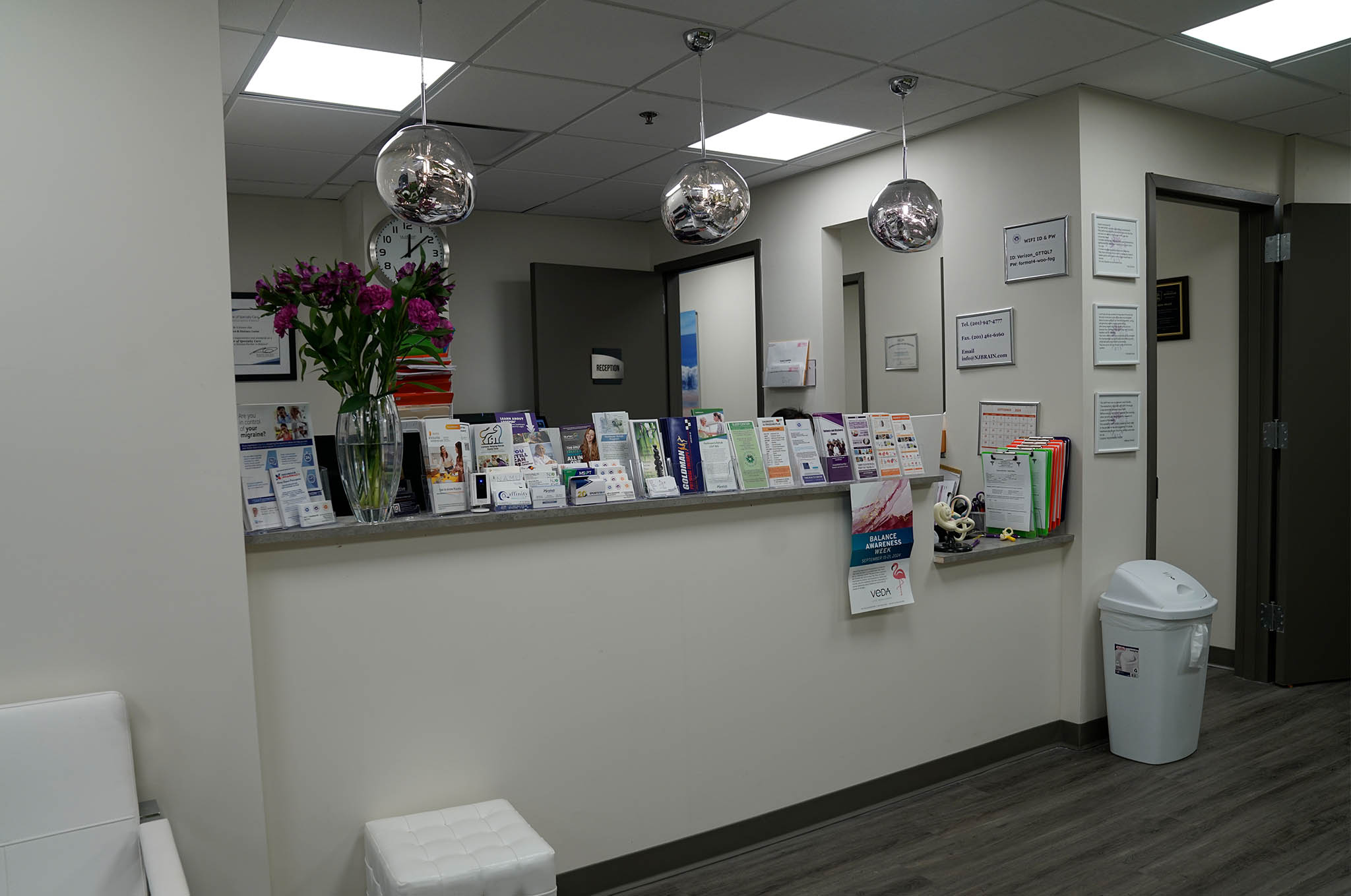 The image shows an interior space that appears to be a waiting area or reception desk within a medical facility. There are posters on the wall, a counter with various items, and a decorative arrangement of spherical lights above the counter.