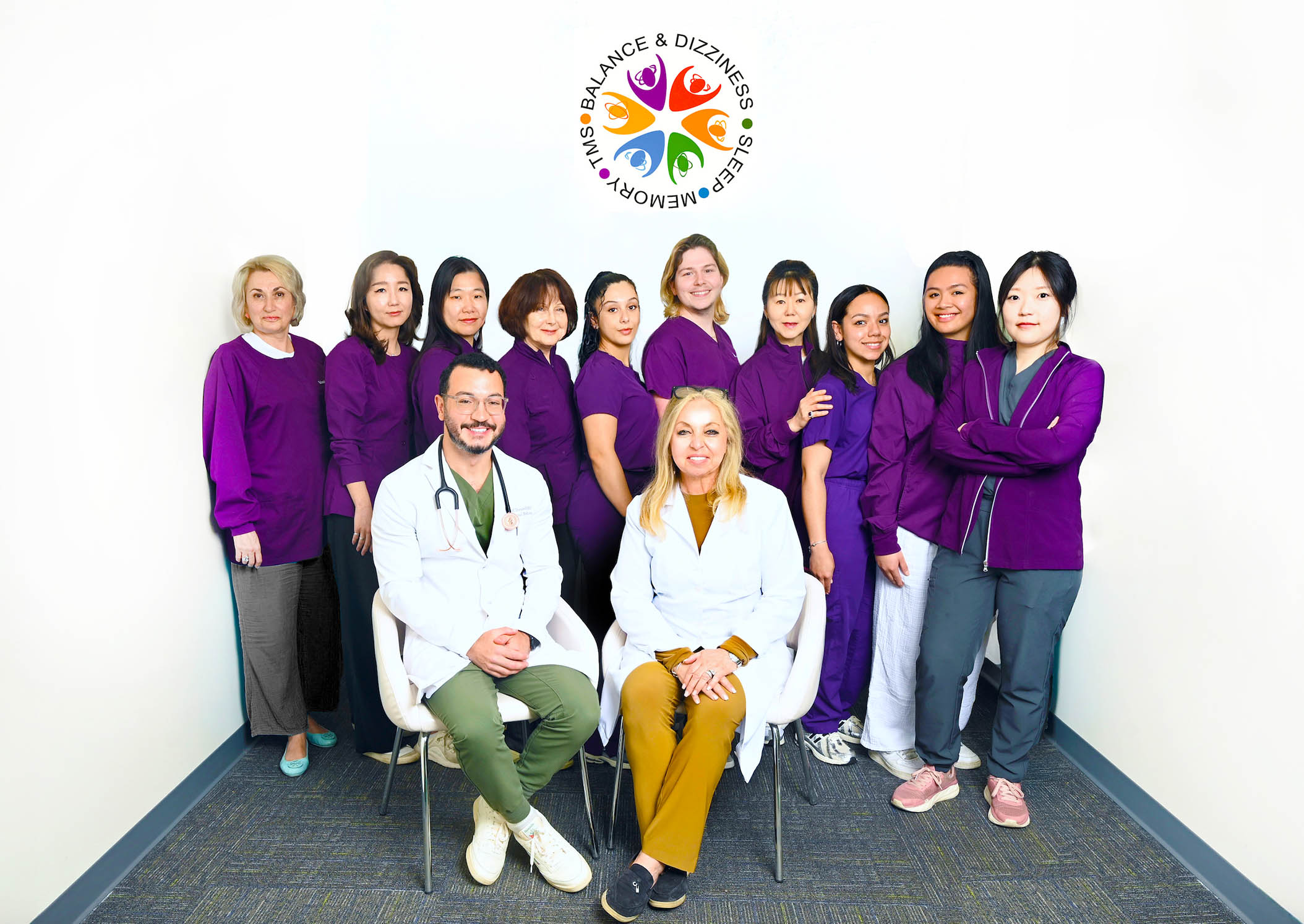 The image shows a group of individuals posing together for a photograph, with some seated and others standing, all dressed in various shades of blue and purple attire, likely representing uniforms for healthcare professionals. They are gathered around a central figure who appears to be an older male wearing a white shirt and stethoscope, seated on the floor between two individuals. The setting is indoors with a wall-mounted plaque featuring text and logos visible behind them.