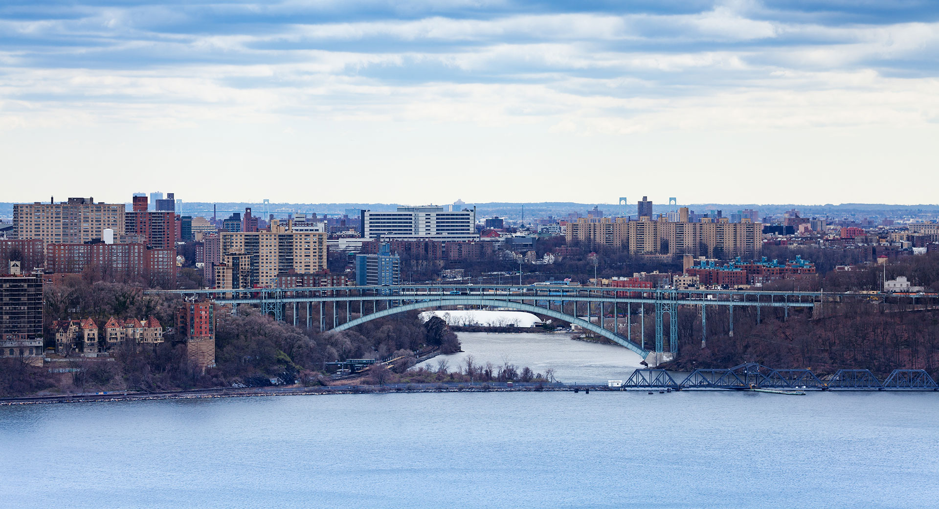 The image displays a panoramic view of a cityscape with a prominent bridge spanning over water, set against a cloudy sky with a few scattered clouds, and features buildings along the riverbank.
