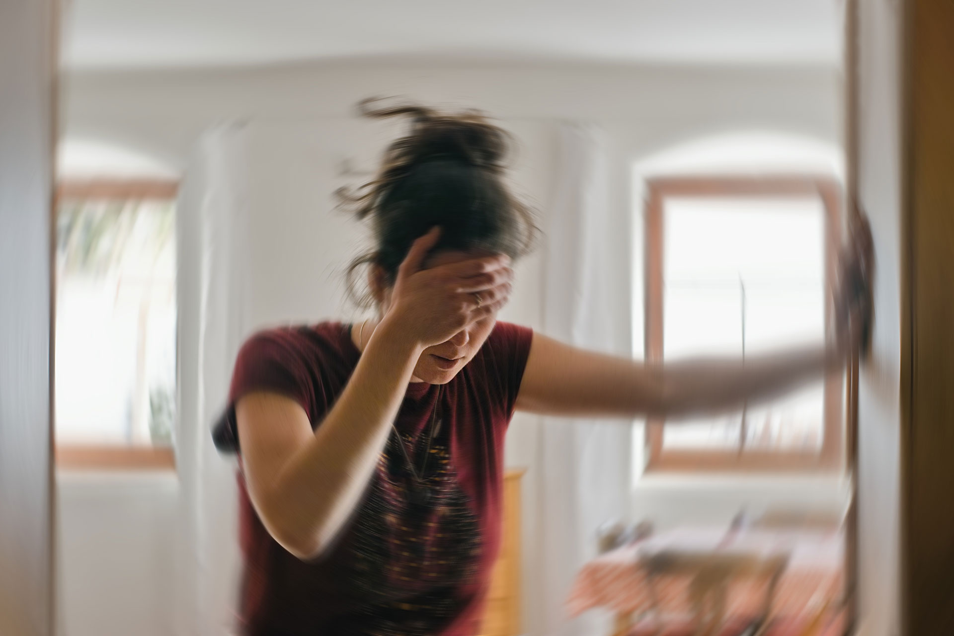 A woman with her hand on her face is captured in motion, creating a blurred effect, while standing in an indoor room with a window.