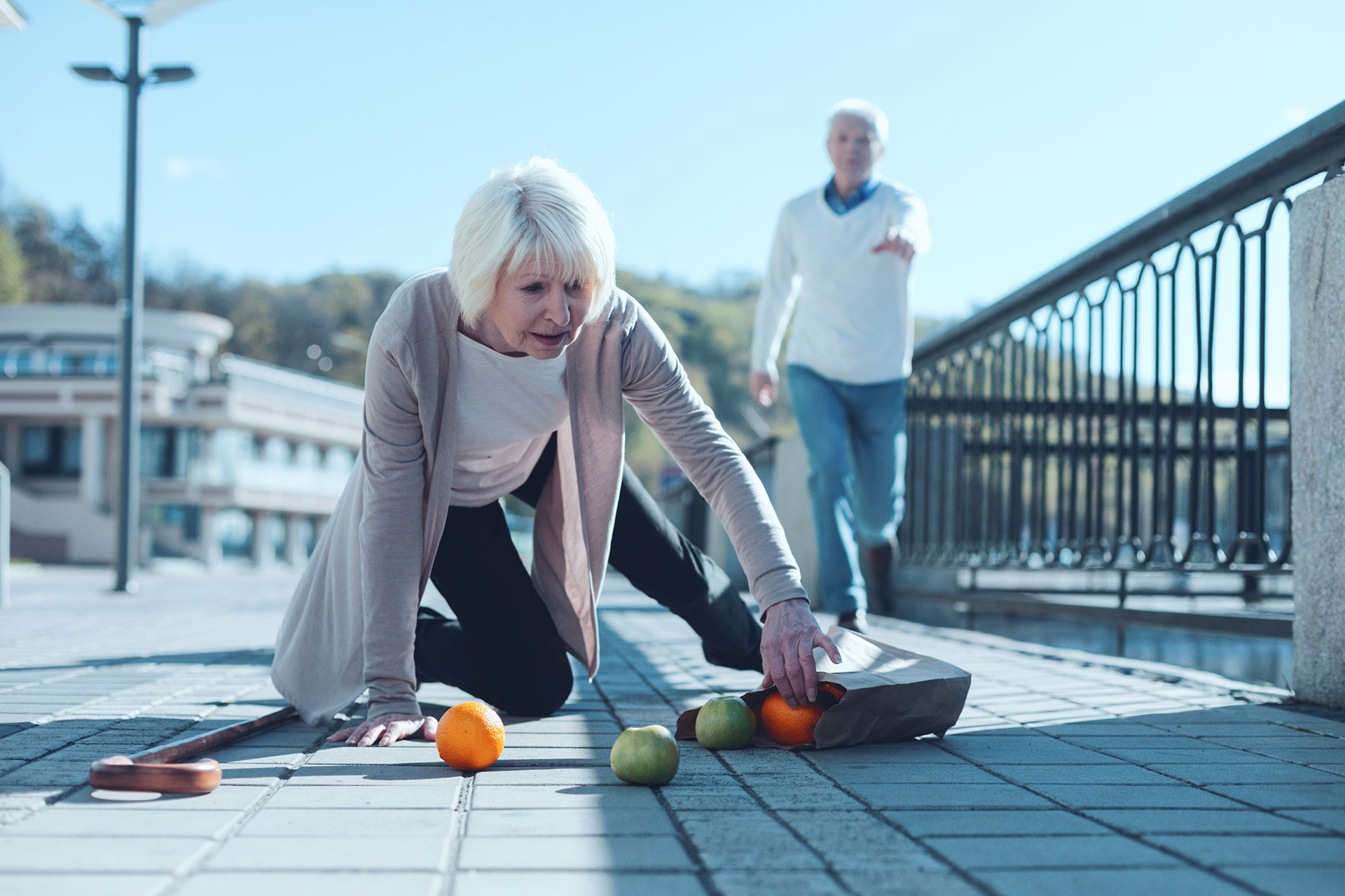 A senior woman bending over to pick up fruit on a sidewalk.