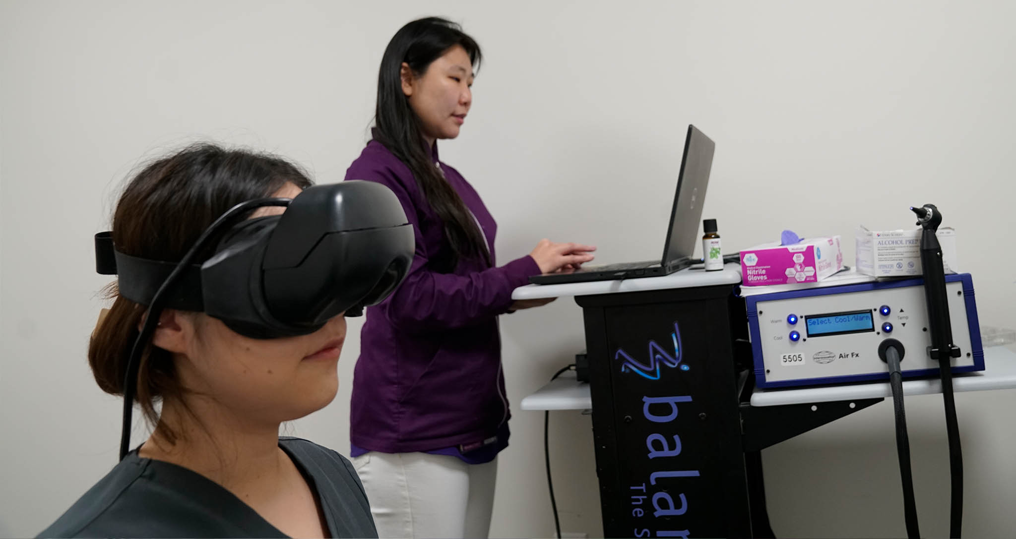 The image shows two individuals engaged with technology in a room; on the left, there's a person wearing a VR headset, and on the right, another individual appears to be operating a device with a laptop and a stethoscope.