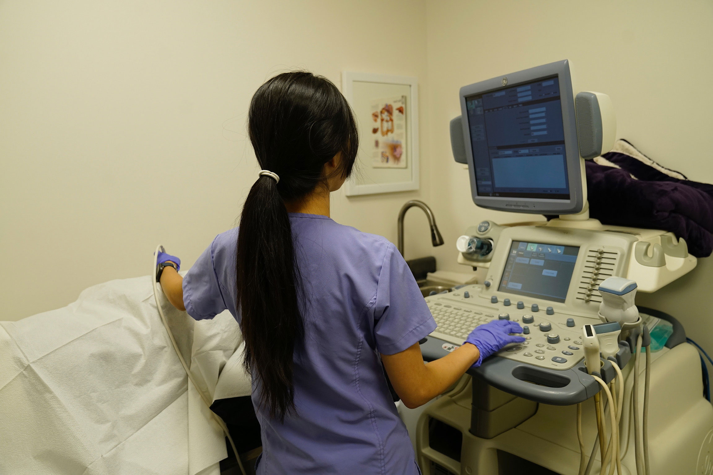 A woman in scrubs stands next to an ultrasound machine, monitoring a patient's scan on a monitor.