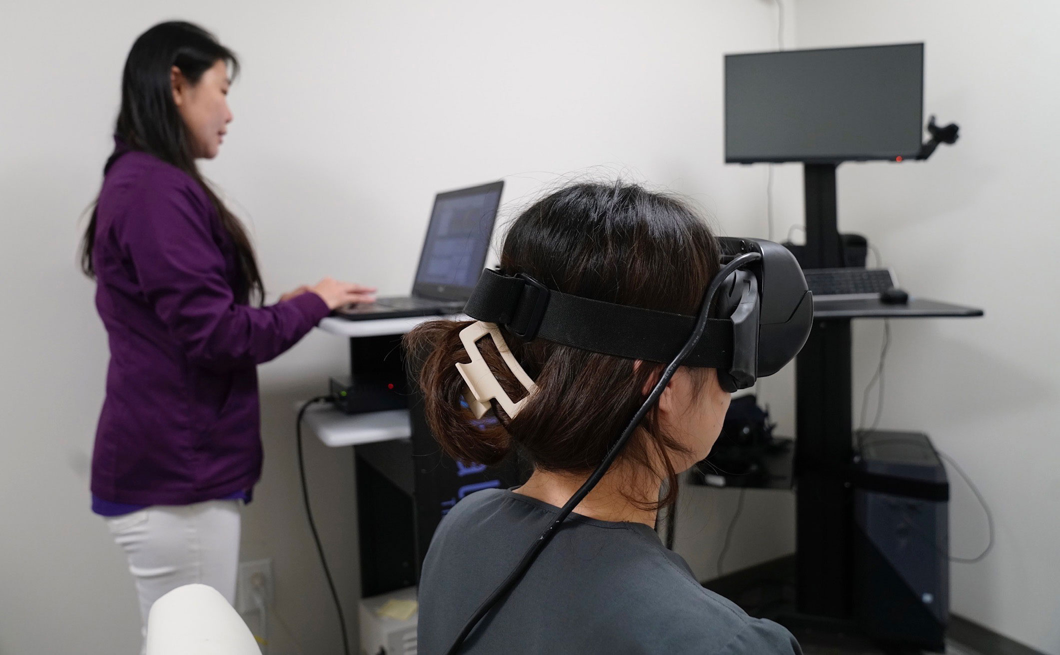 In the image, there are two individuals engaged with technology: one person is standing at a workstation using a laptop, while another person is seated in front of a monitor wearing a virtual reality headset. The scene suggests a workspace where technology is being utilized for various purposes.