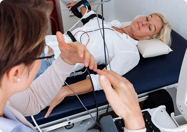 A woman lying on a medical examination table with an attached heart rate monitor, being attended to by a healthcare professional who appears to be adjusting the equipment.