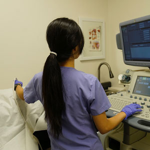 A woman with long hair wearing scrubs stands at an ultrasound machine in a hospital room.